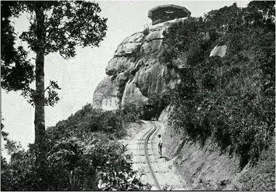 Train entering a tunnel beneath a large rock outcrop, tracks winding through the forest, historic photo